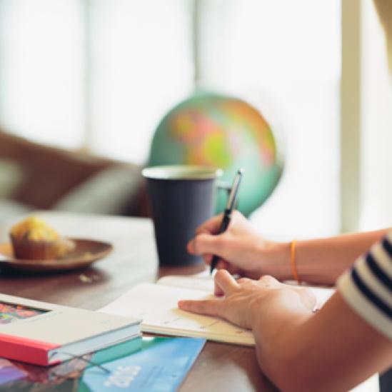 A person taking notes at the table with books, coffee, and a muffin placed on it